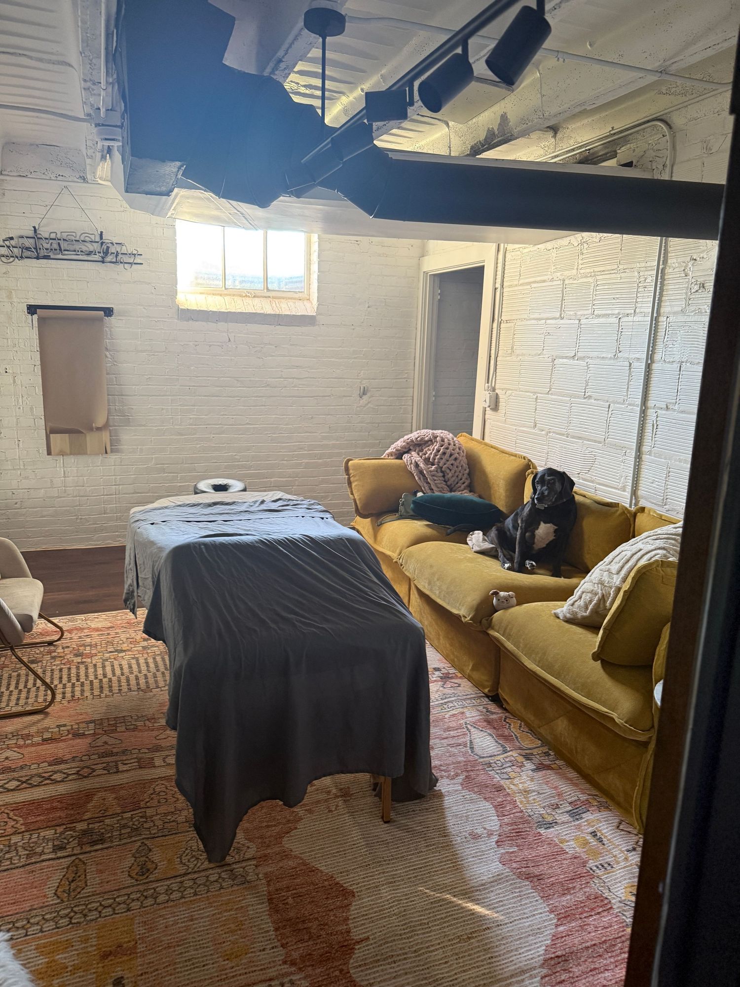 Modern basement lounge area with yellow velvet sofa, oriental rug, and exposed ceiling beams in industrial style space.