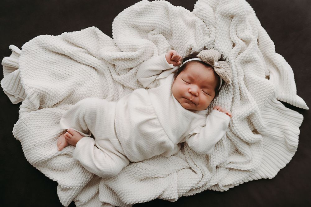 A sleeping newborn baby dressed in white lies peacefully on a textured cream-colored knit blanket.