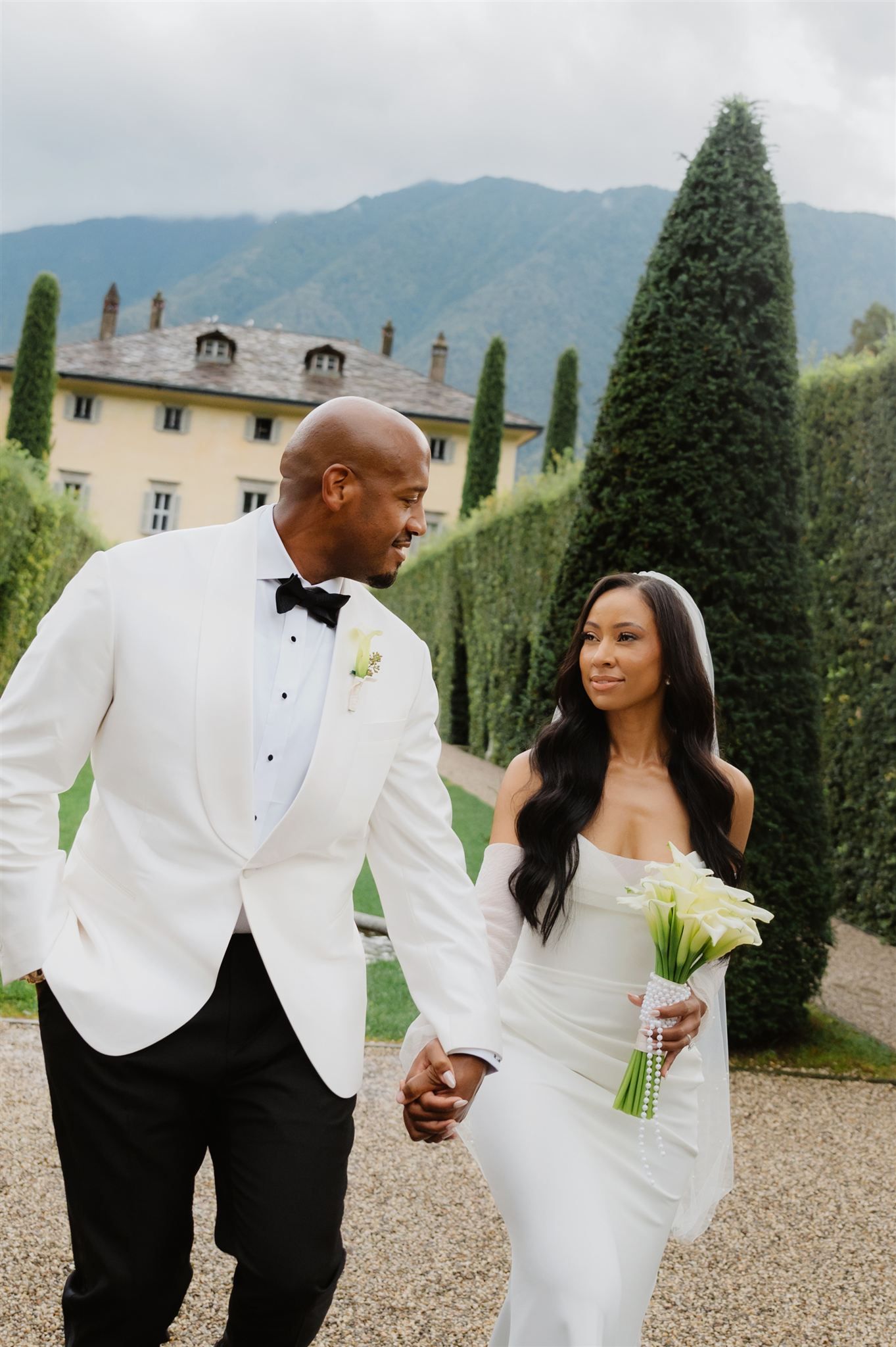 Wedding couple in white attire posing in garden with mountain backdrop and cypress trees.