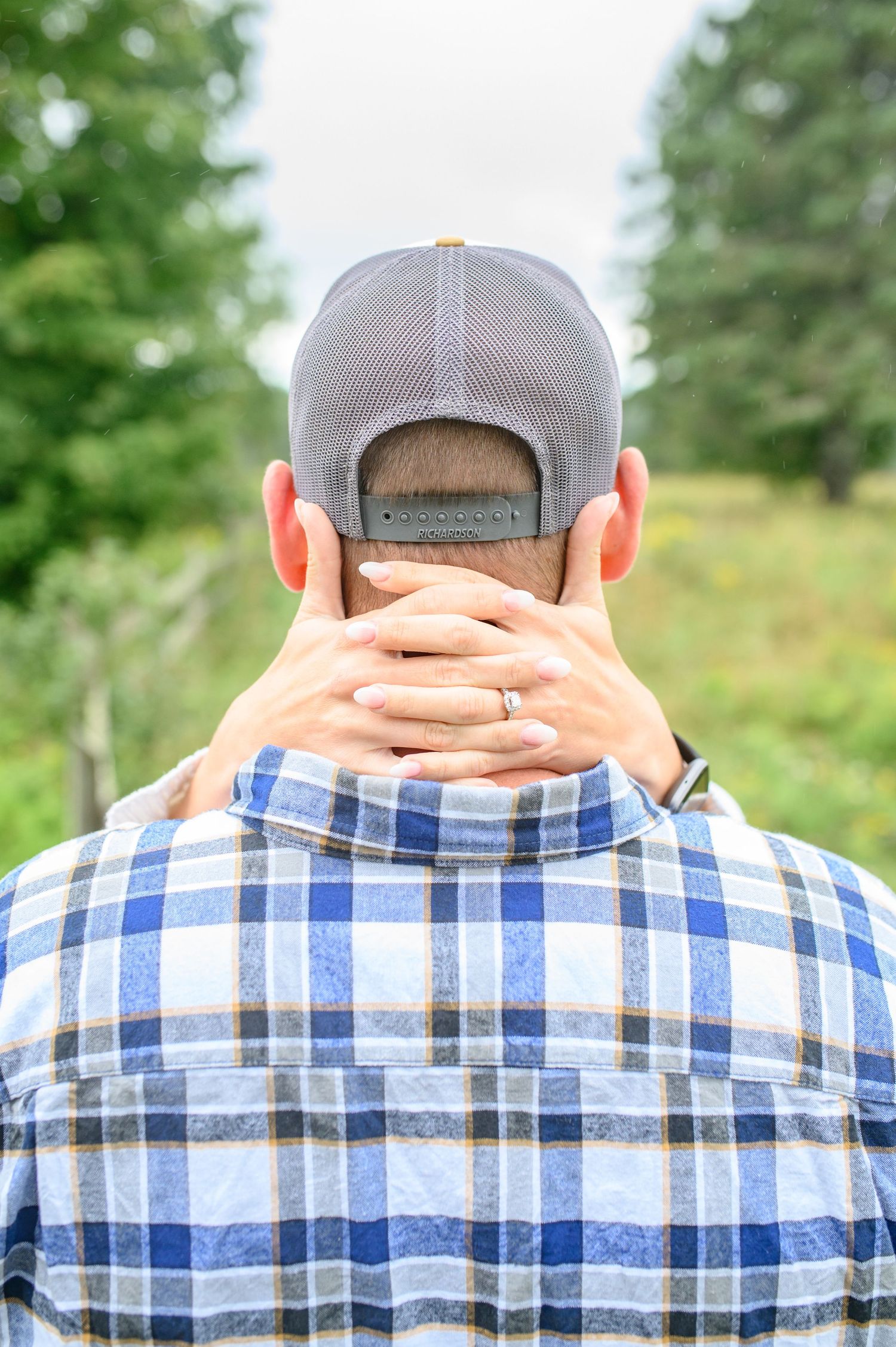 Back view of person wearing gray baseball cap and blue plaid shirt against green outdoor background.