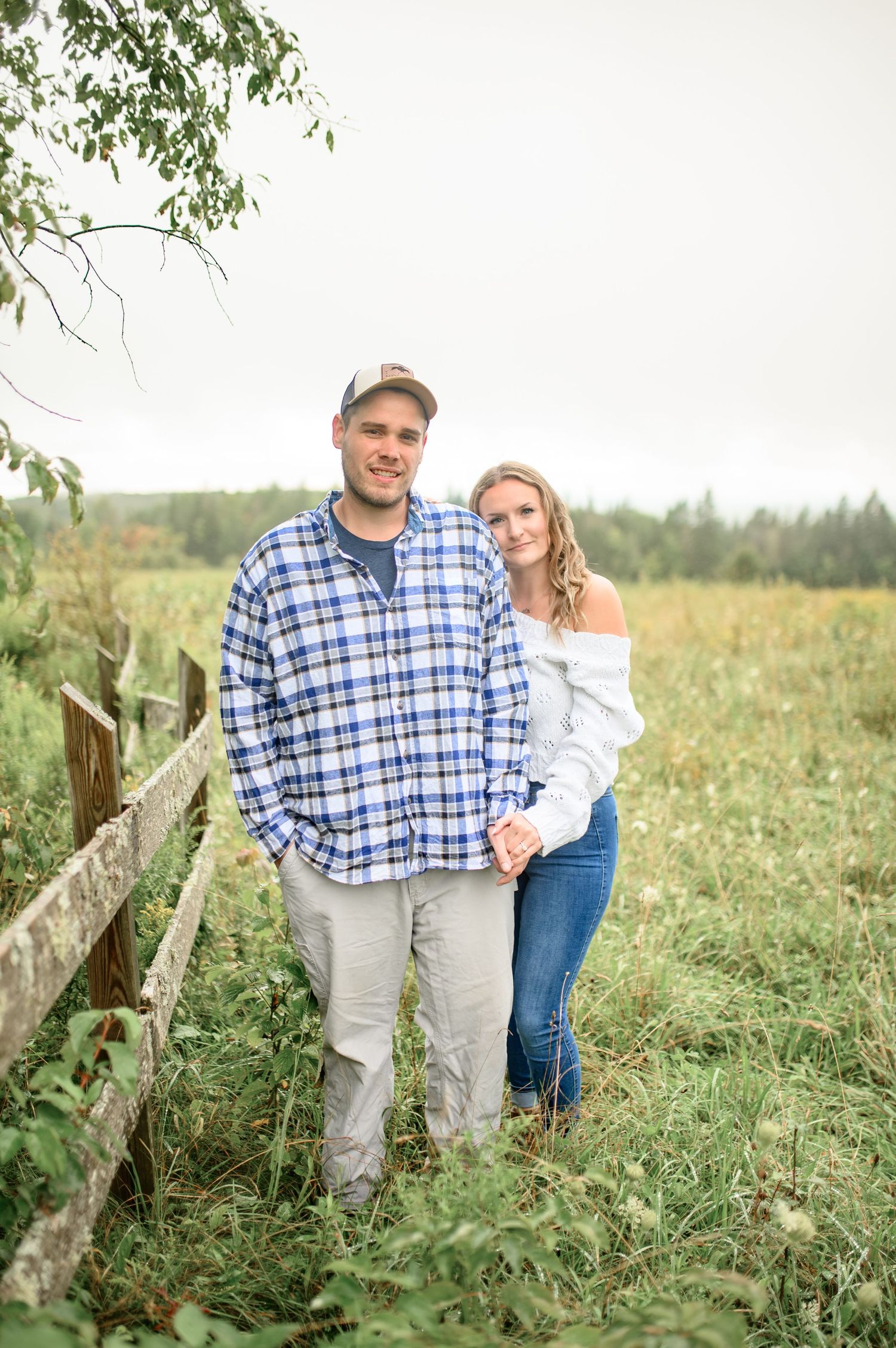 Couple standing together in field with rustic fence and greenery on overcast day.