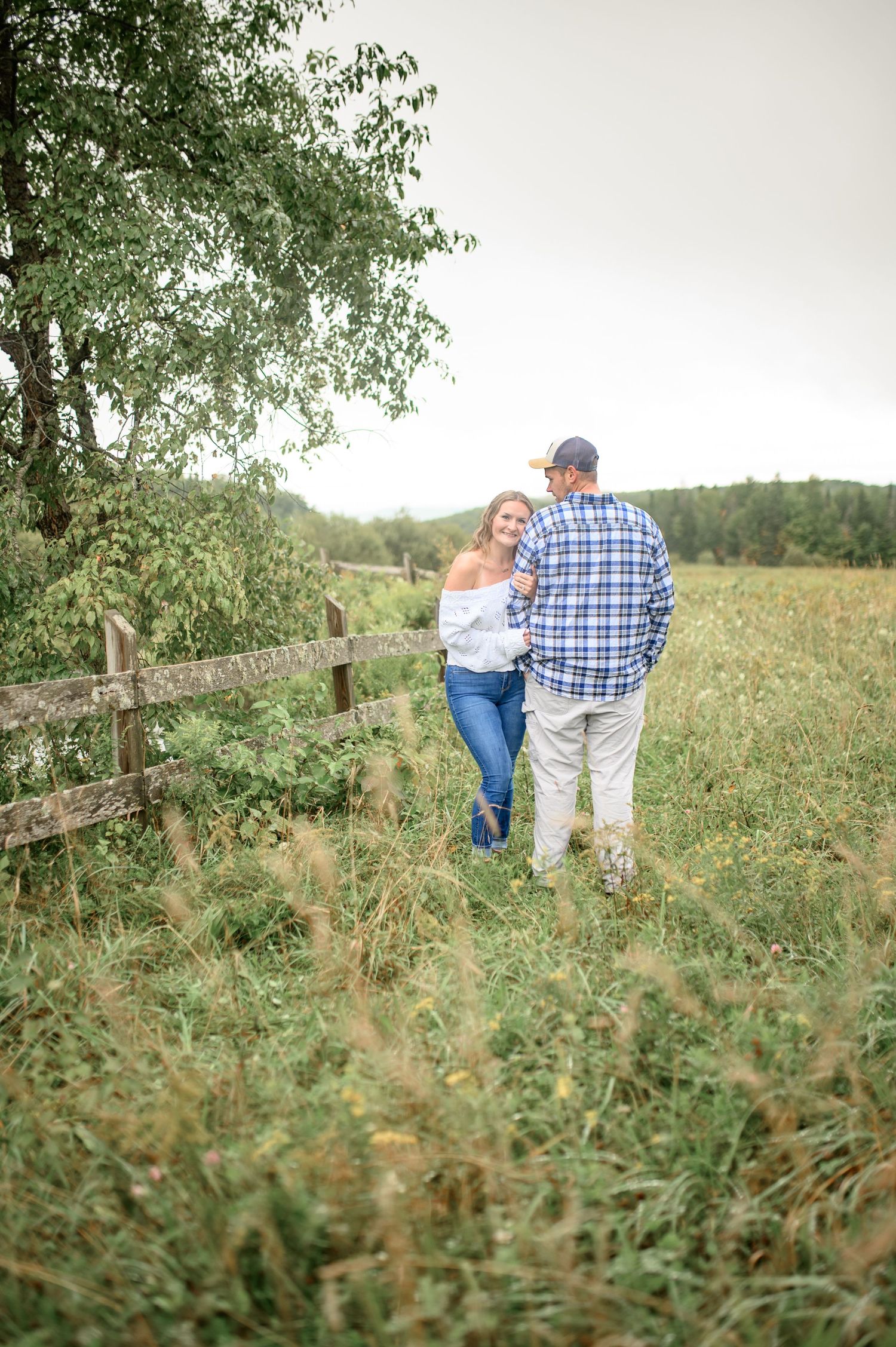 A couple stands together among wildflowers and grass in a serene country setting with overcast skies.