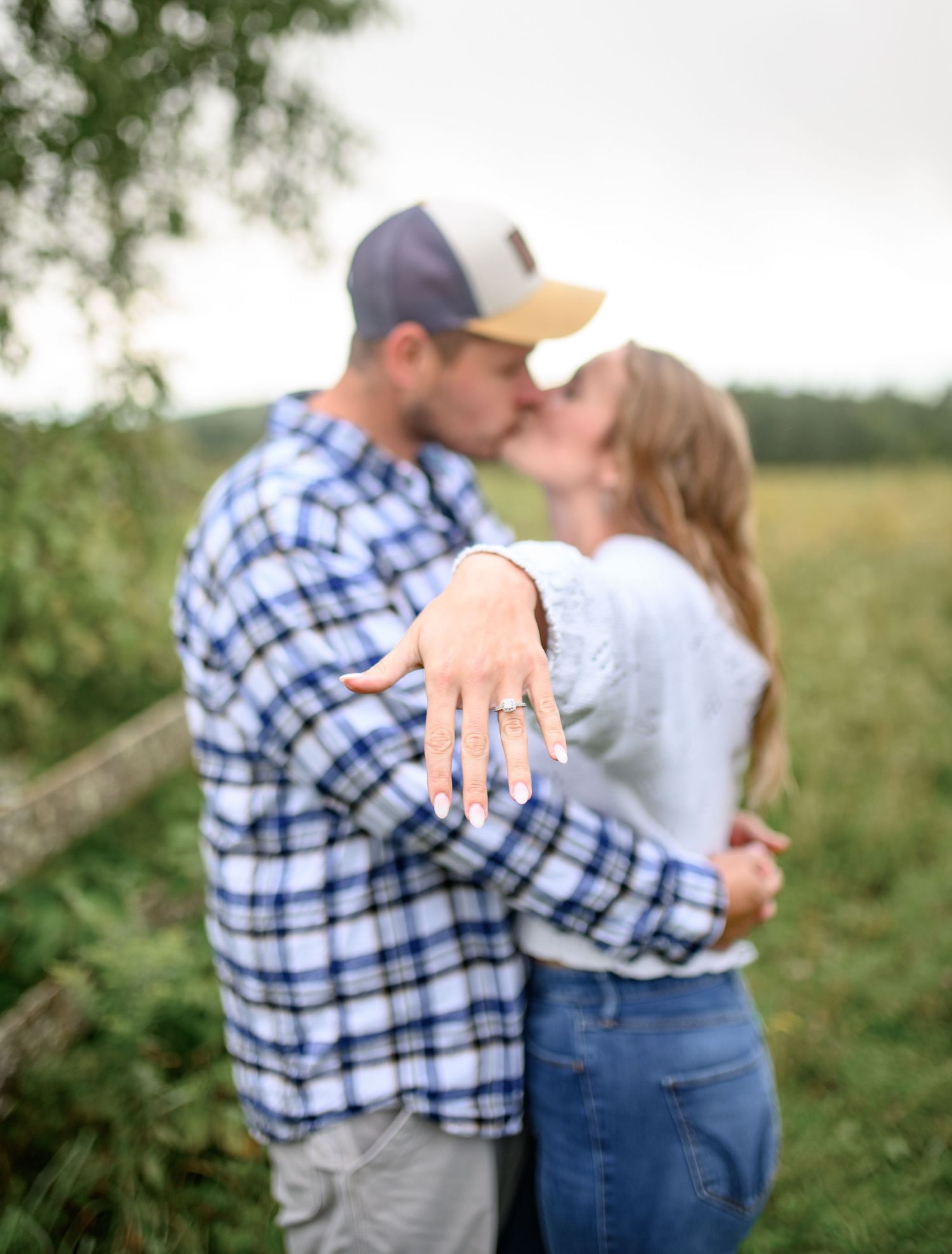 A couple embraces in an outdoor field with green foliage while showing off an engagement ring.