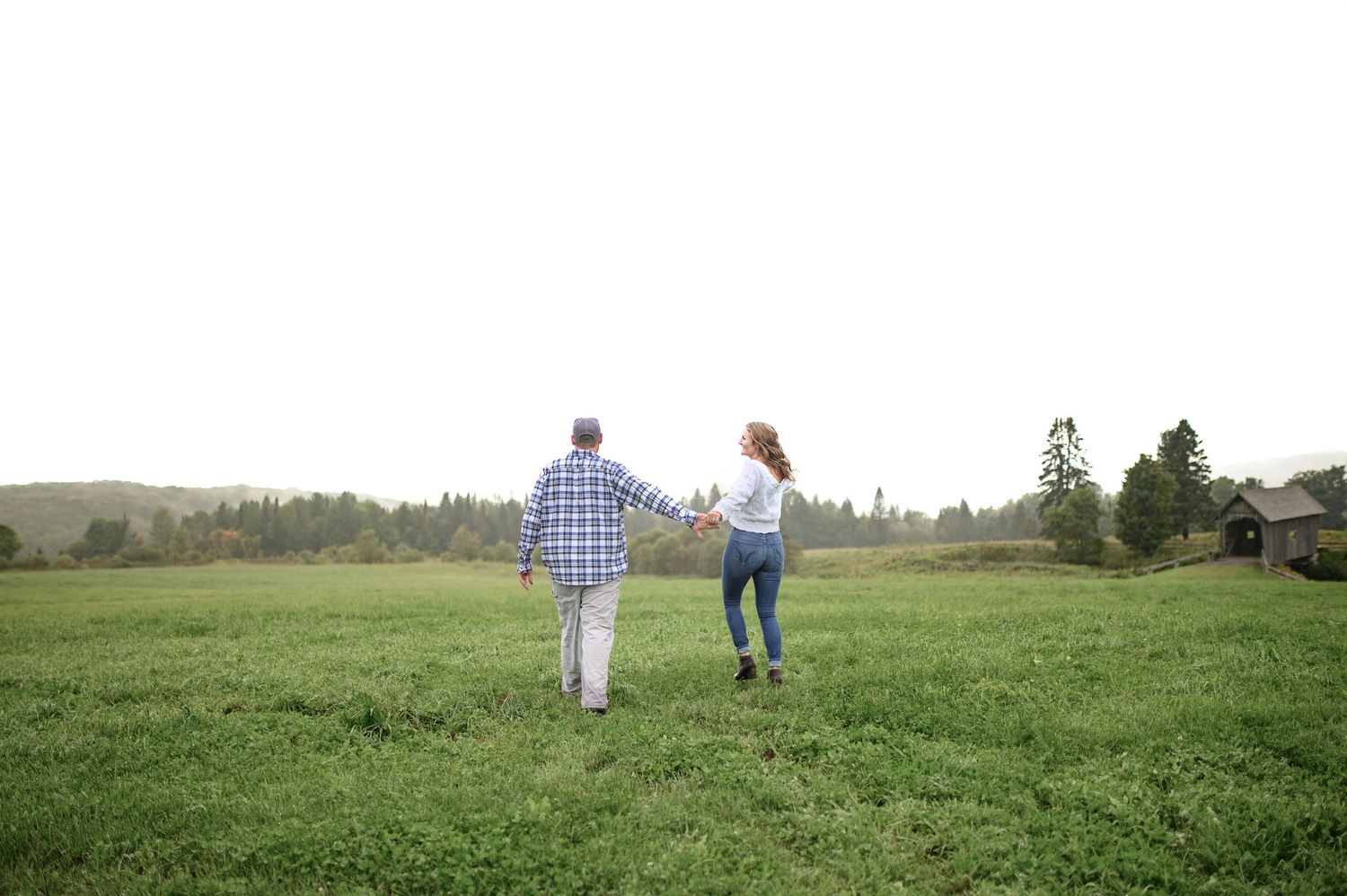A couple walks hand in hand across a lush green meadow with mountains and trees in the background.