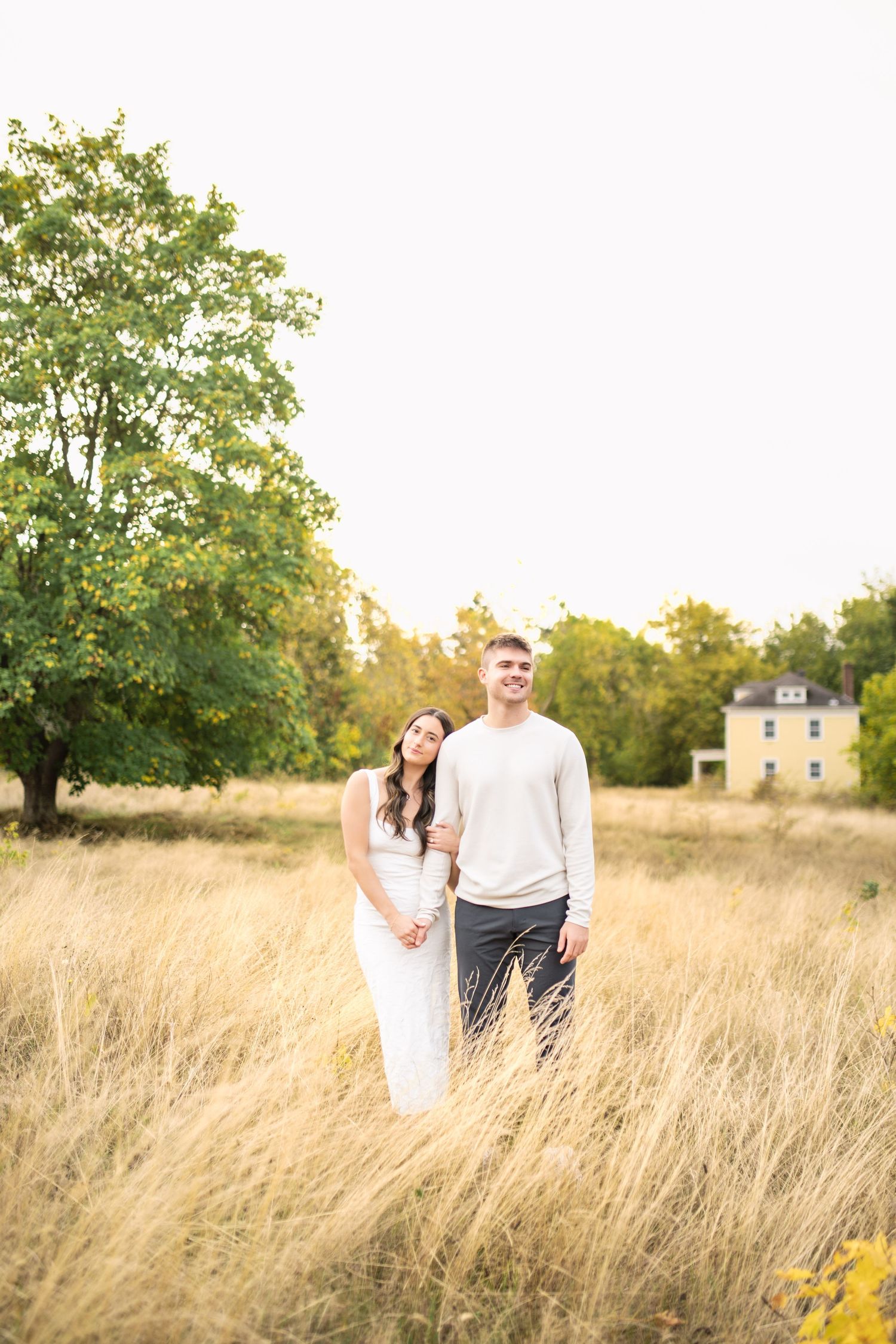 A couple stands together in a golden field of tall grass during sunset with trees in the background.