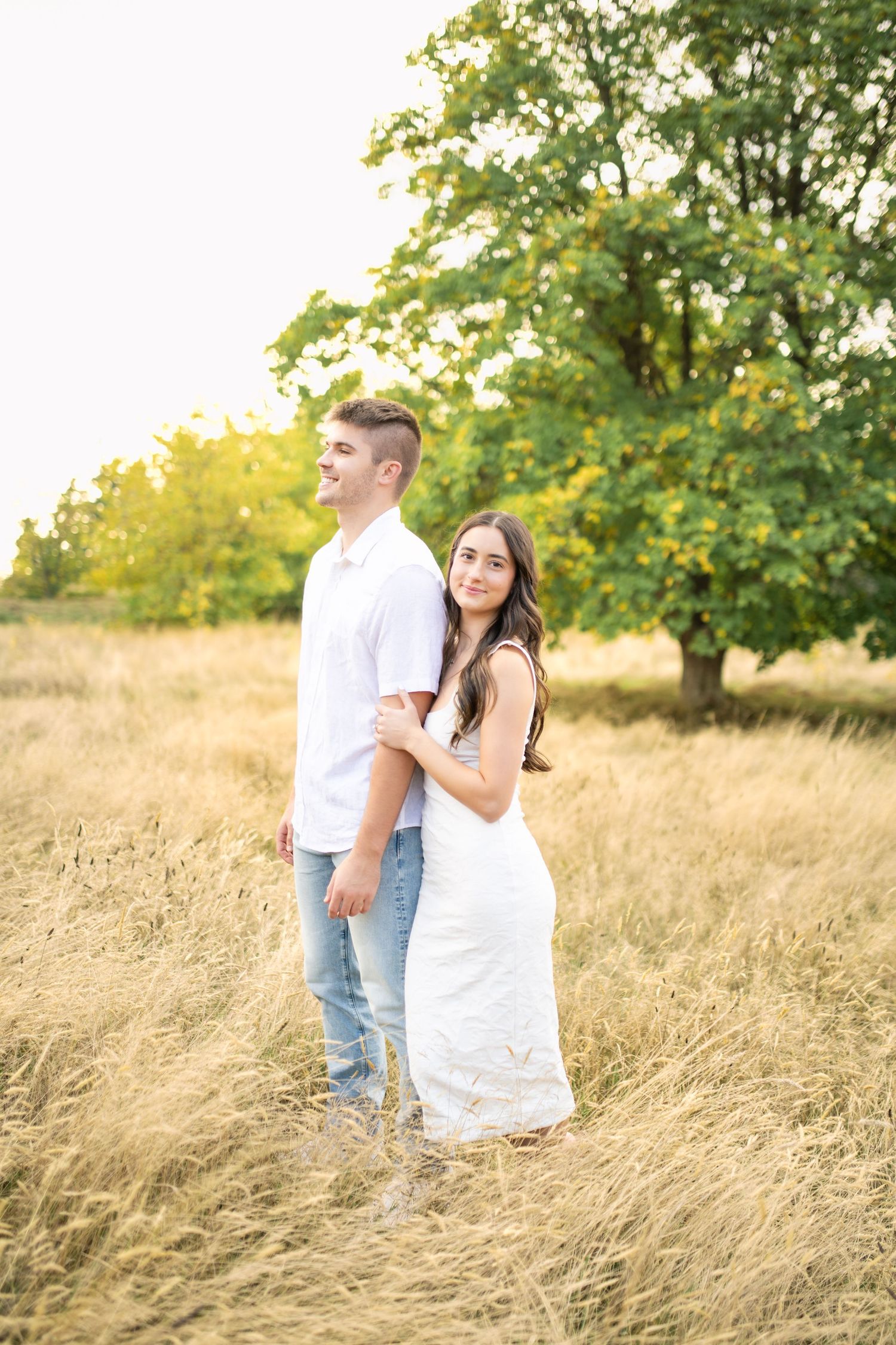 A couple poses together in a golden field at sunset with trees in the background.