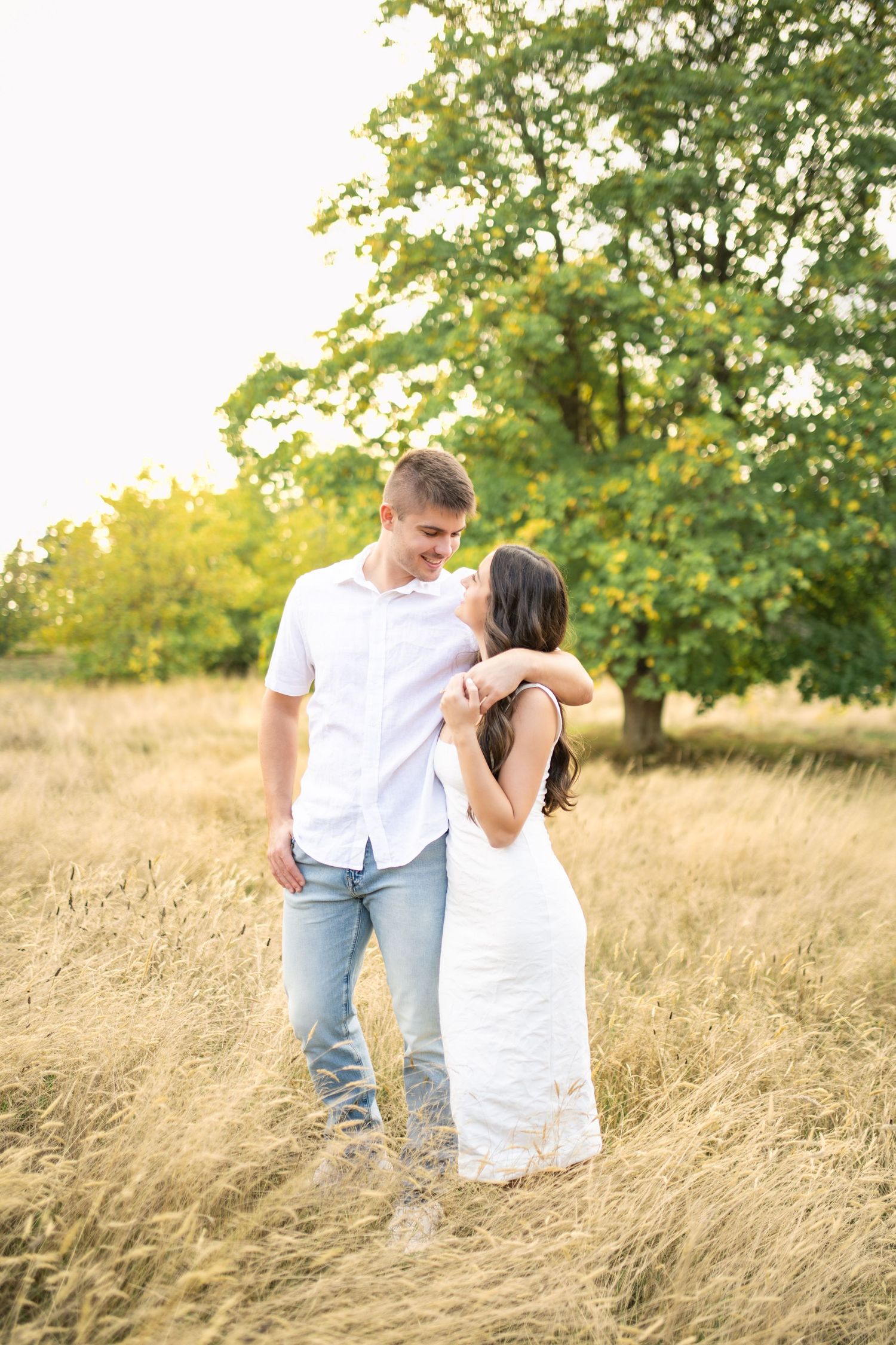 A romantic couple in white clothing embraces while standing in a golden wheat field during sunset.