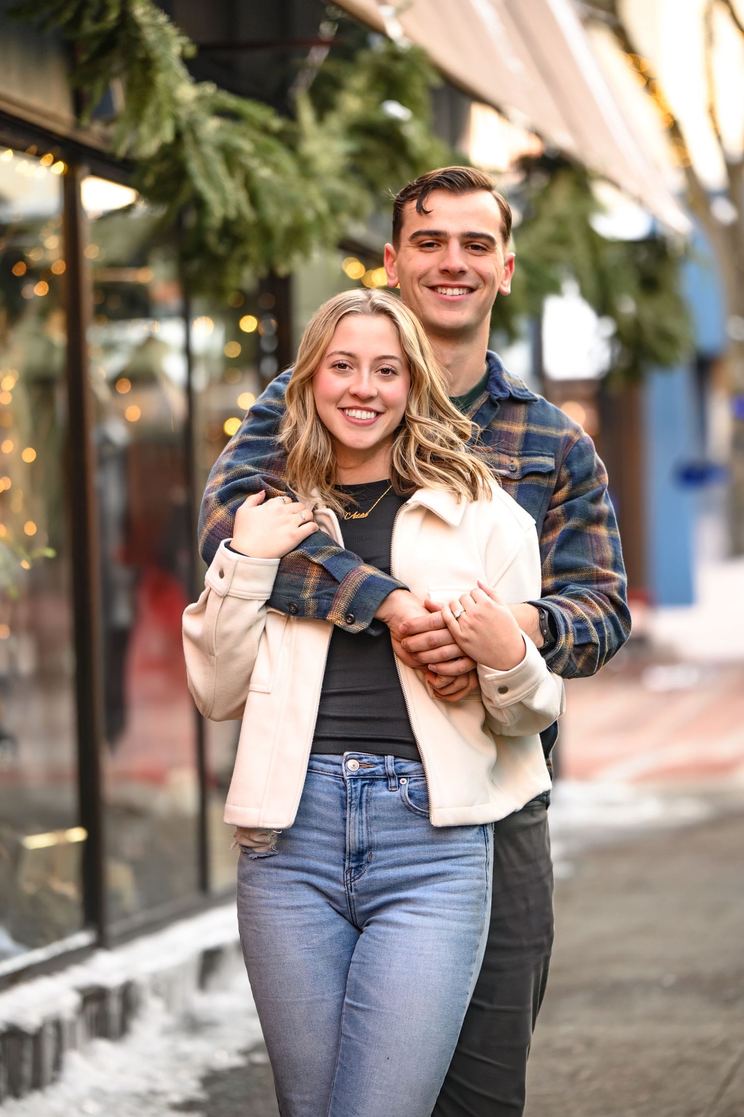 Couple laughing and embracing while walking along a city sidewalk with palm trees and storefronts in background.