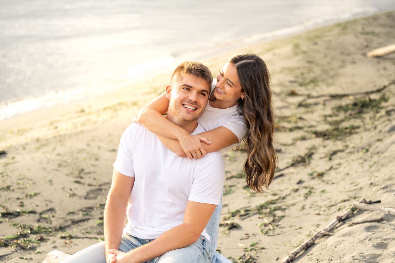 A couple in white outfits shares intimate moments during a romantic beach photoshoot at sunset.