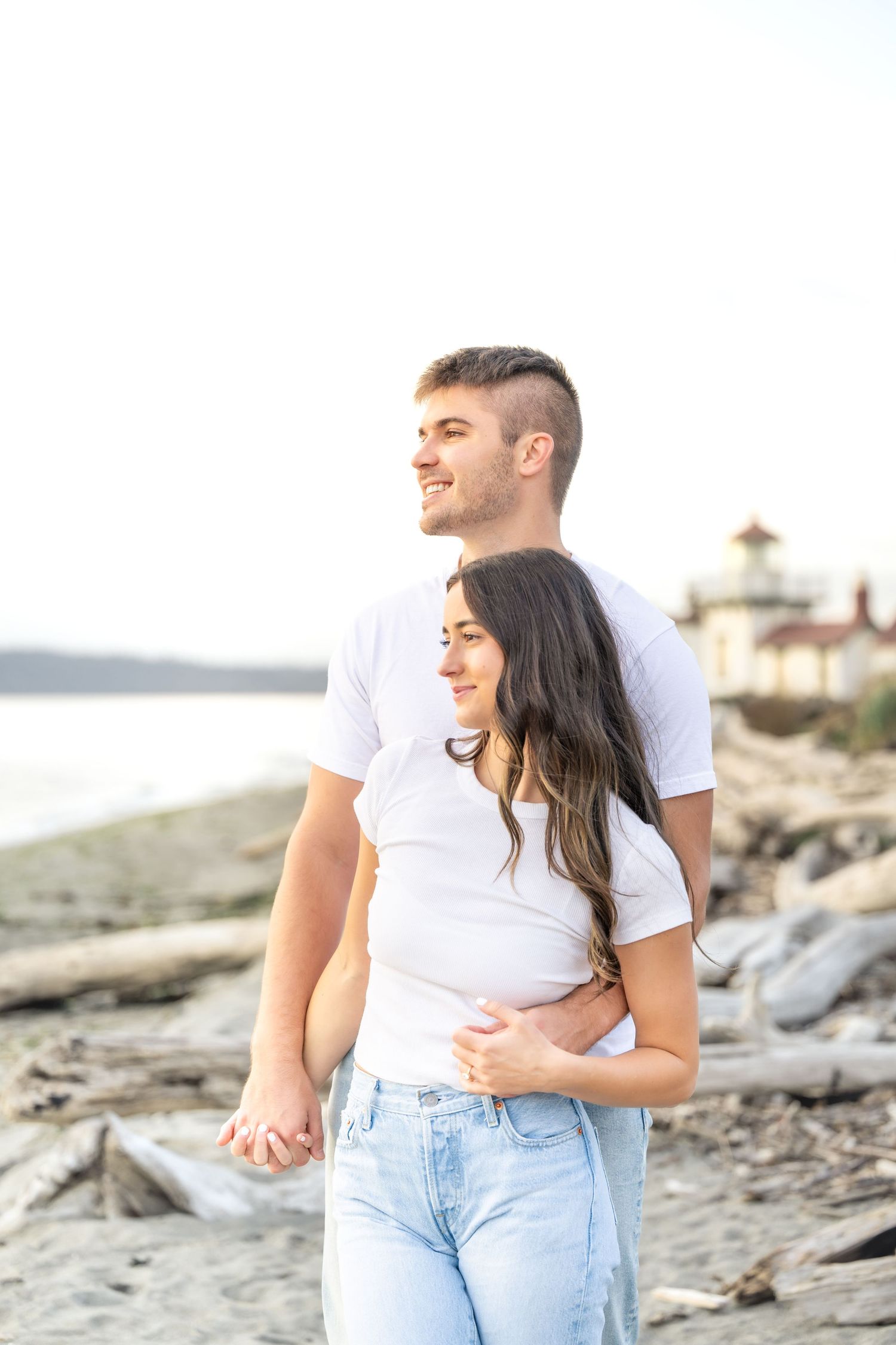 Romantic couple walking together on a rocky beach shore in casual white and denim outfits.