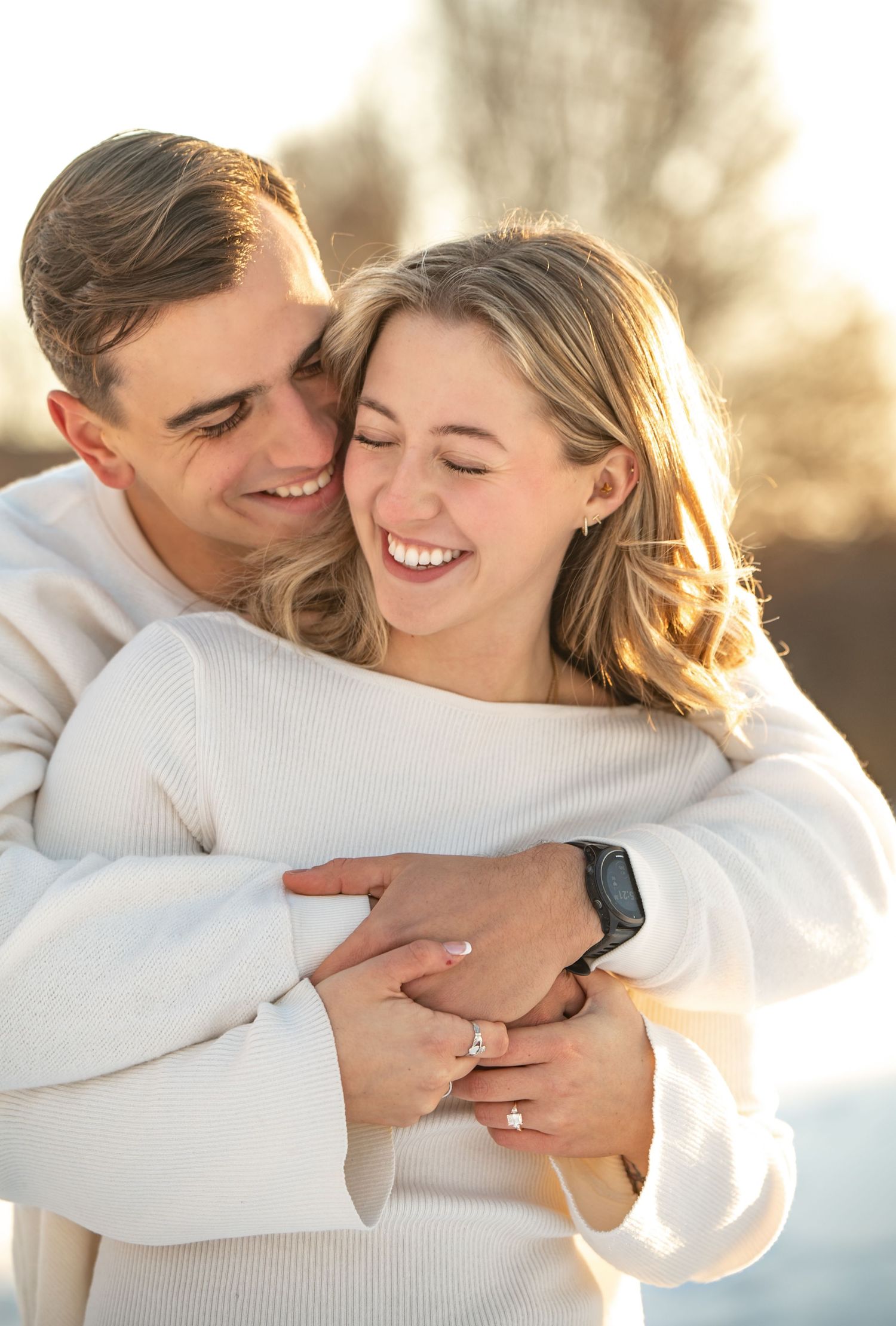 A couple embraces in warm sunlight wearing white sweaters while sharing a joyful intimate moment together.