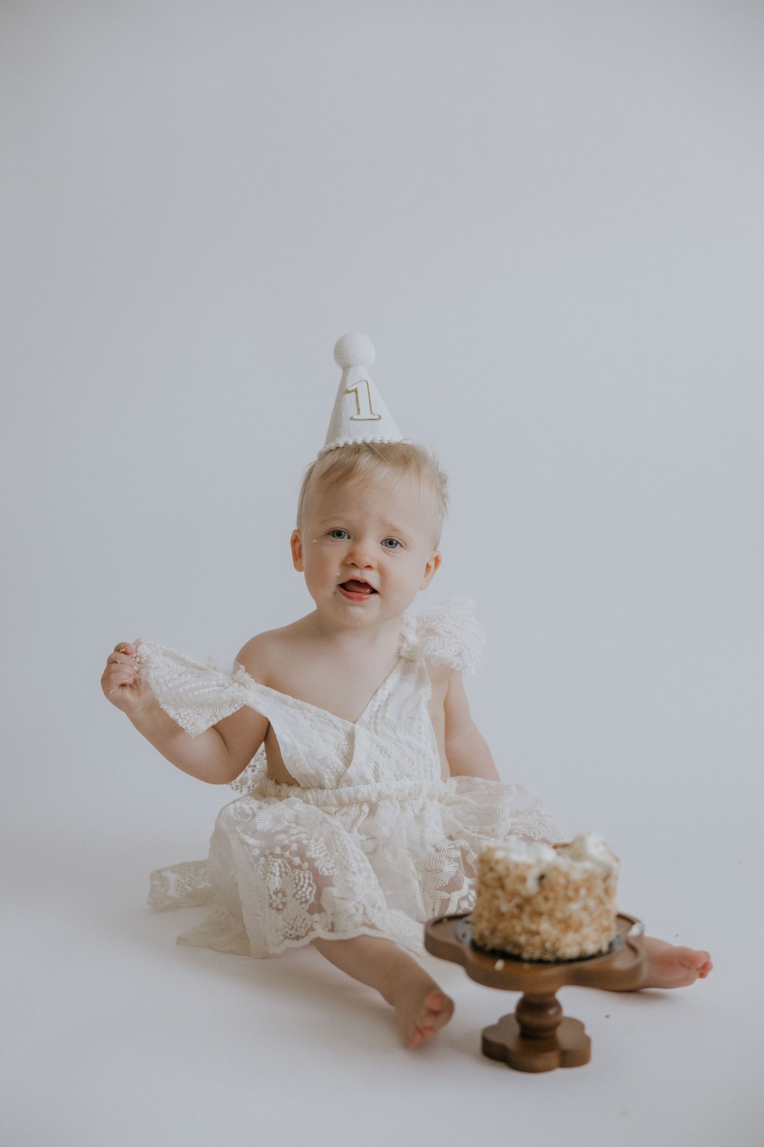 Baby in white lace dress sits on floor with cake and cookies against light gray background.
