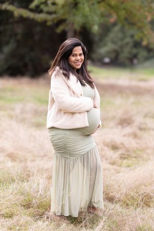 A person in a cream sweater and sage green dress poses gracefully in a sunlit meadow with tall grass.