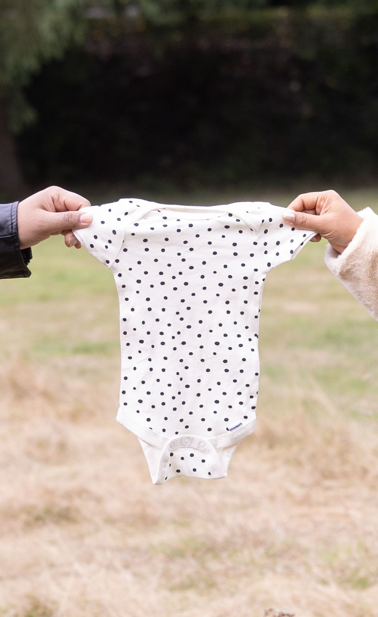 Hands hold up a white polka dot baby onesie against a blurred outdoor background.