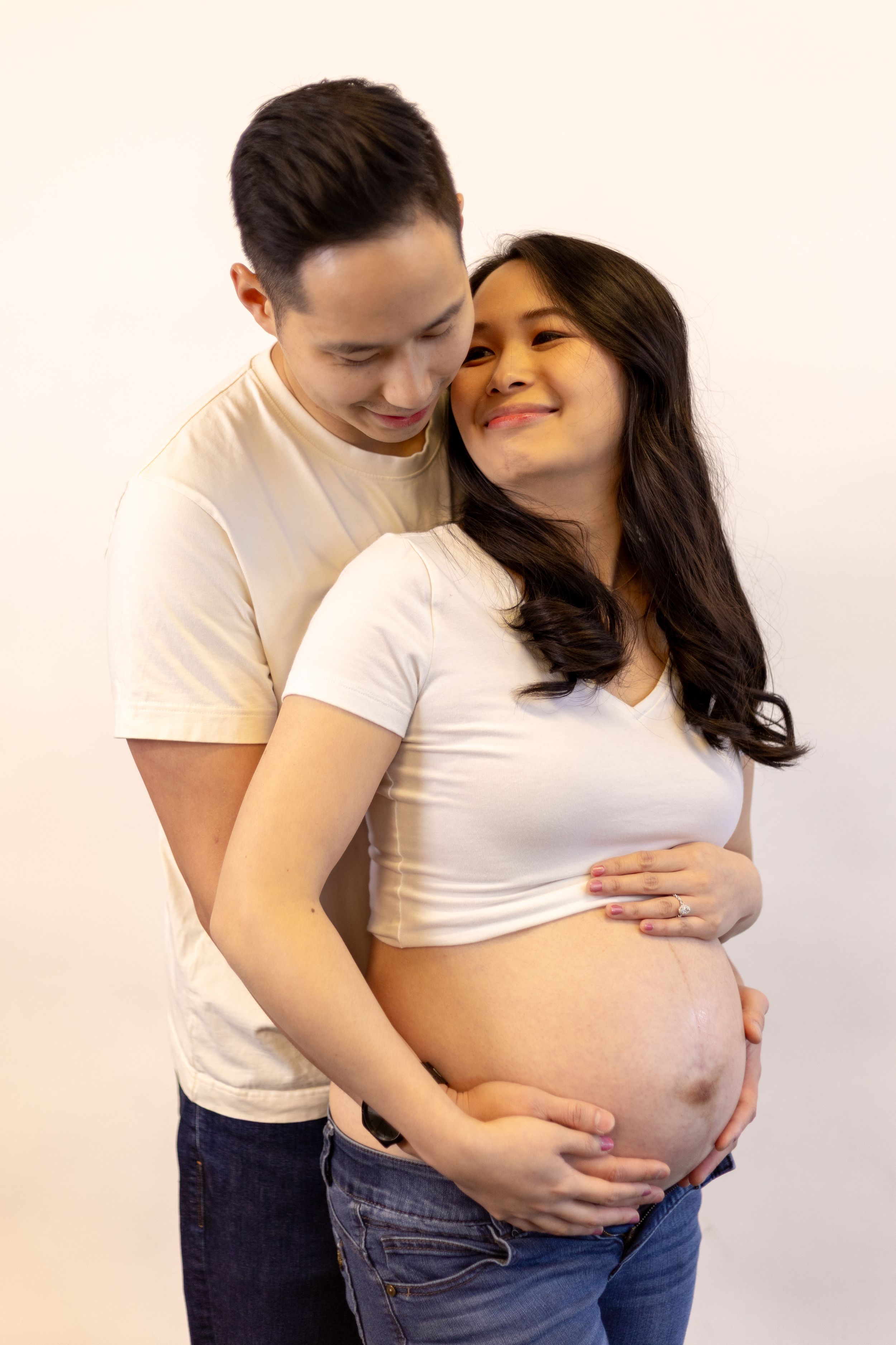 A couple shares an intimate moment during a maternity photoshoot against a white backdrop.