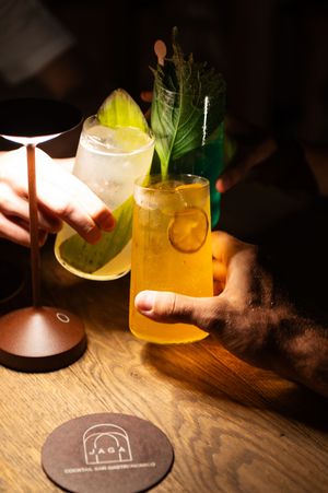 Cocktails with citrus garnishes on wooden table with branded coasters in dim lighting.
