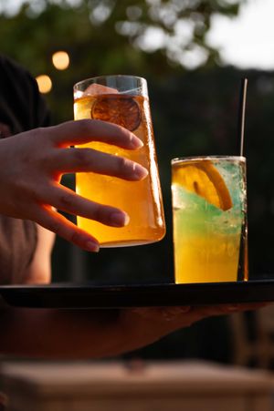 Hands reaching for colorful cocktails garnished with citrus in evening lighting.