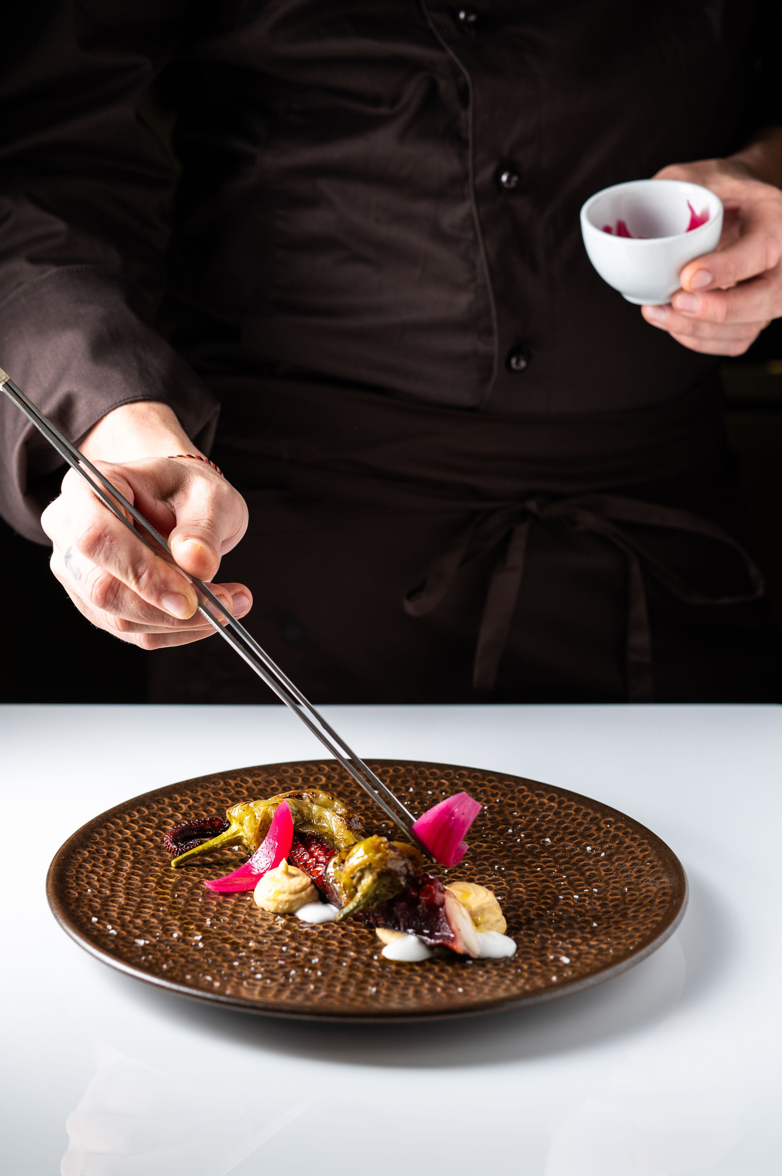 Chef plating gourmet dish with chopsticks on textured brown plate, garnished with pink flowers and greens.