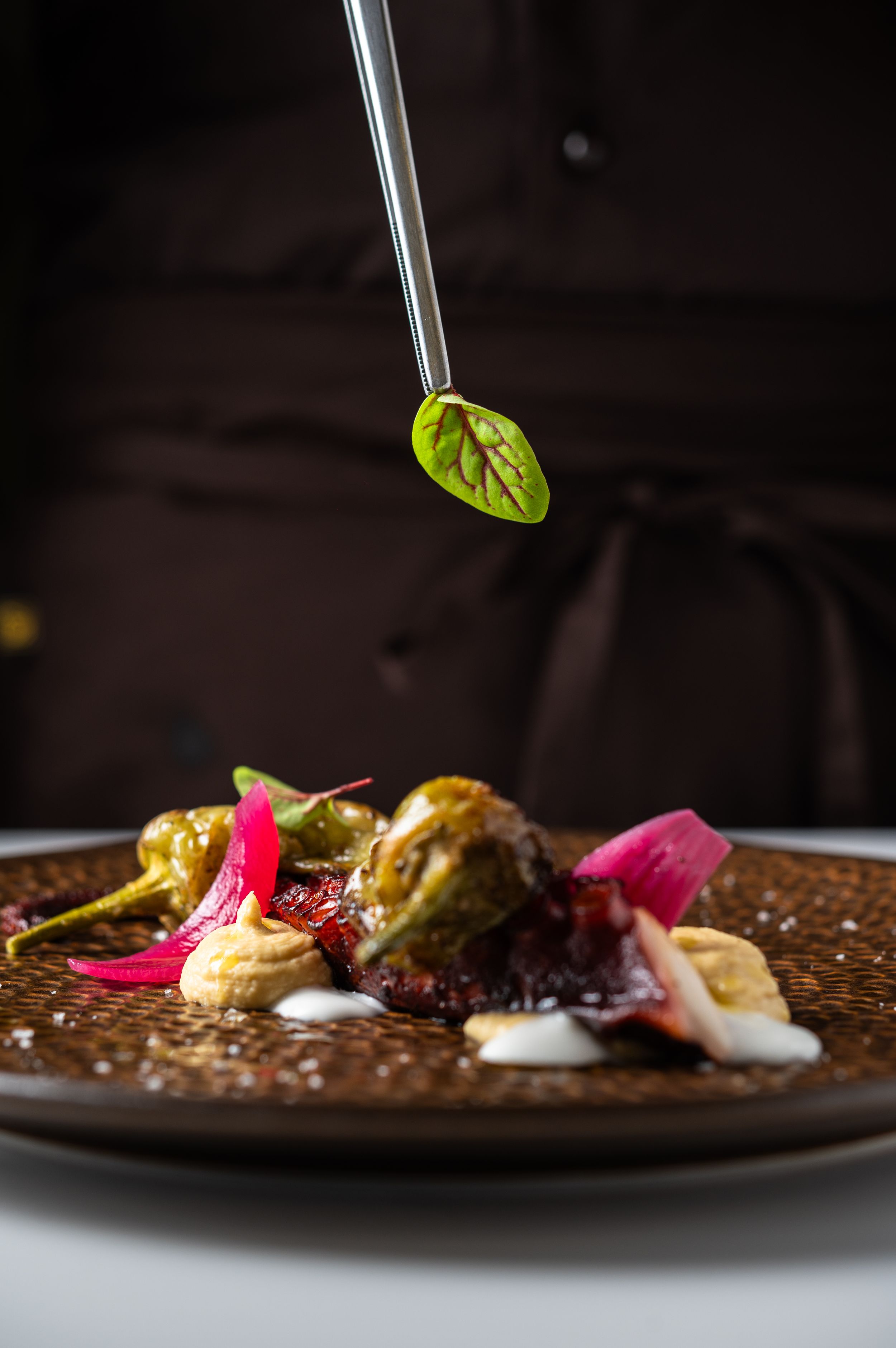 Chef using tweezers to place a fresh green leaf on an elegant gourmet dish with colorful garnishes on a brown plate.