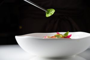 Chef placing fresh basil leaf on elegant white bowl with colorful garnishes in fine dining presentation.