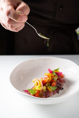Chef drizzling green herb oil over a colorful gourmet tuna tartare dish in a white bowl.