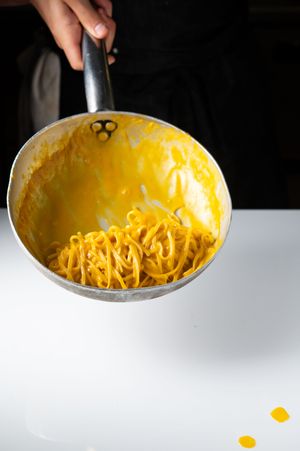 Chef tossing fresh yellow spaghetti pasta in a silver saucepan with creamy egg sauce.