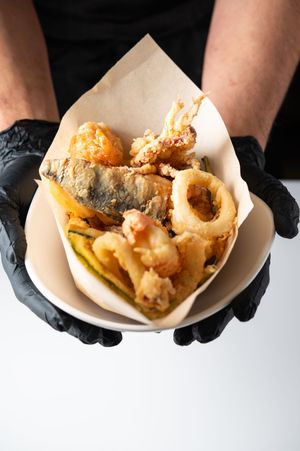 Hands in black gloves holding a paper cone filled with crispy tempura seafood and vegetables.