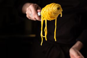 Chef twirling fresh yellow tagliatelle pasta on a fork against a dark background.