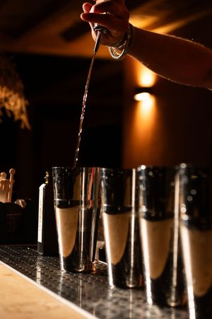 Bartender pouring drinks into multiple metal shakers in low light.
