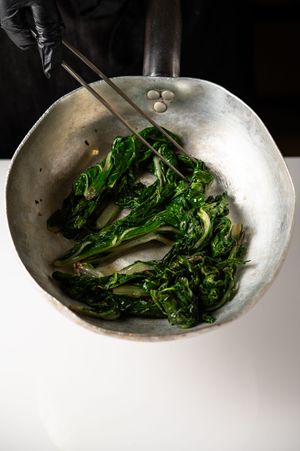 Chef sautéing fresh green vegetables in a worn steel pan with tongs, wearing black gloves.