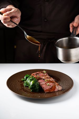 Chef drizzling rich brown sauce over sliced medium-rare beef steak with sautéed spinach on a dark plate.