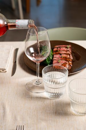 Rose wine being poured into a glass beside a plate of sliced steak and water glasses on a white tablecloth.