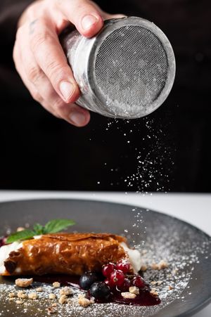 Chef dusting powdered sugar over a golden pastry dessert with blueberries and berry sauce on a gray plate.