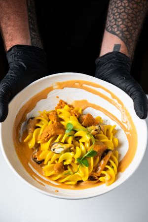 Tattooed hands in black gloves holding a white bowl of fusilli pasta with mussels, sea urchin, and orange bisque sauce.