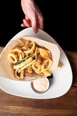 Hand holding a white plate of crispy fried calamari and zucchini with dipping sauce on parchment paper.