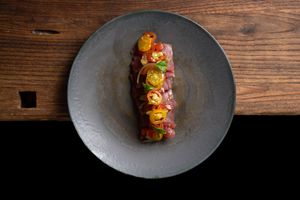 Gourmet beef tartare topped with colorful cherry tomatoes and fresh herbs, elegantly plated on a dark ceramic dish.