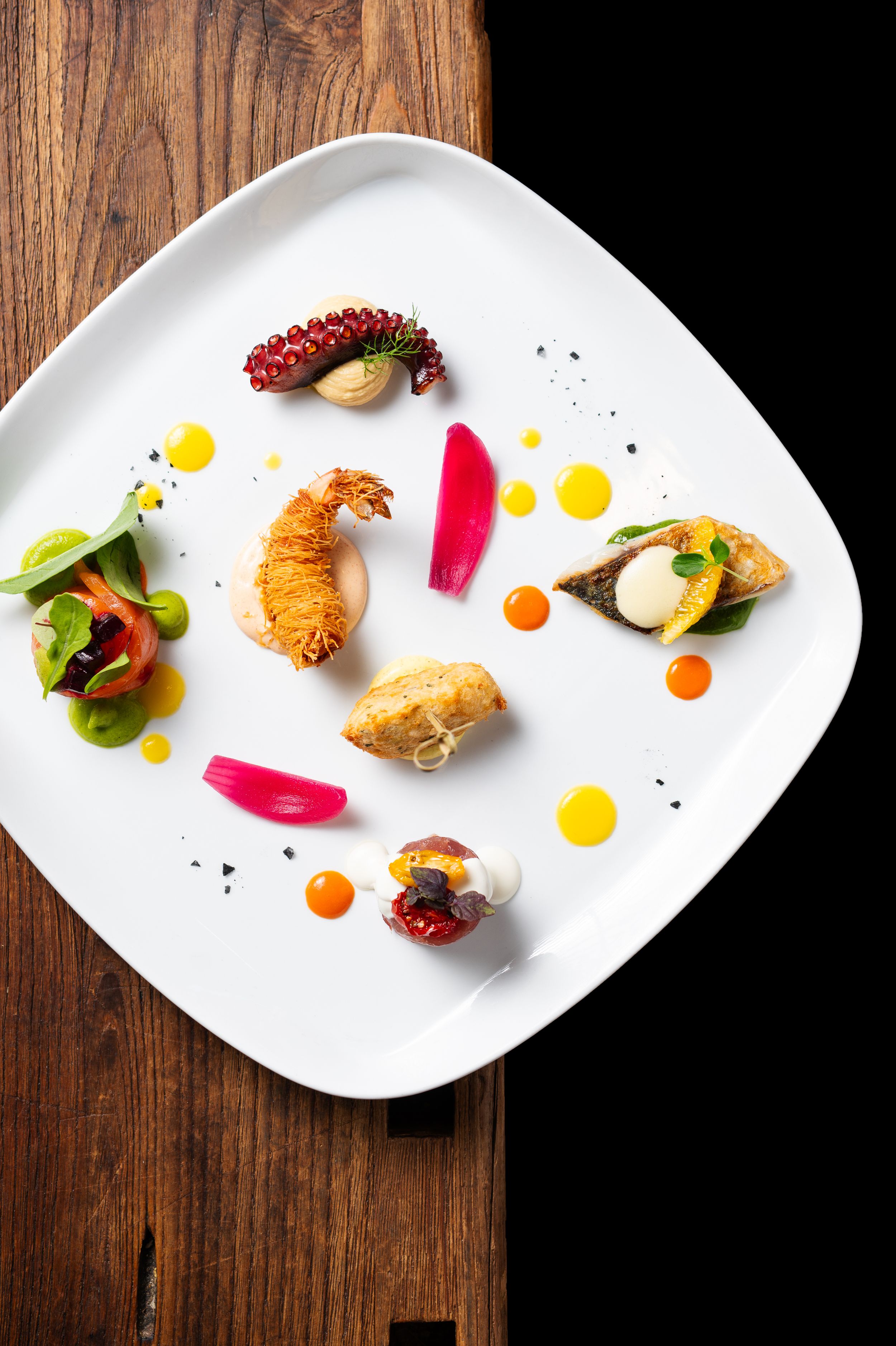 Artistic fine dining seafood tasting plate with crispy prawns, pickled vegetables, and colorful garnishes on white square plate.