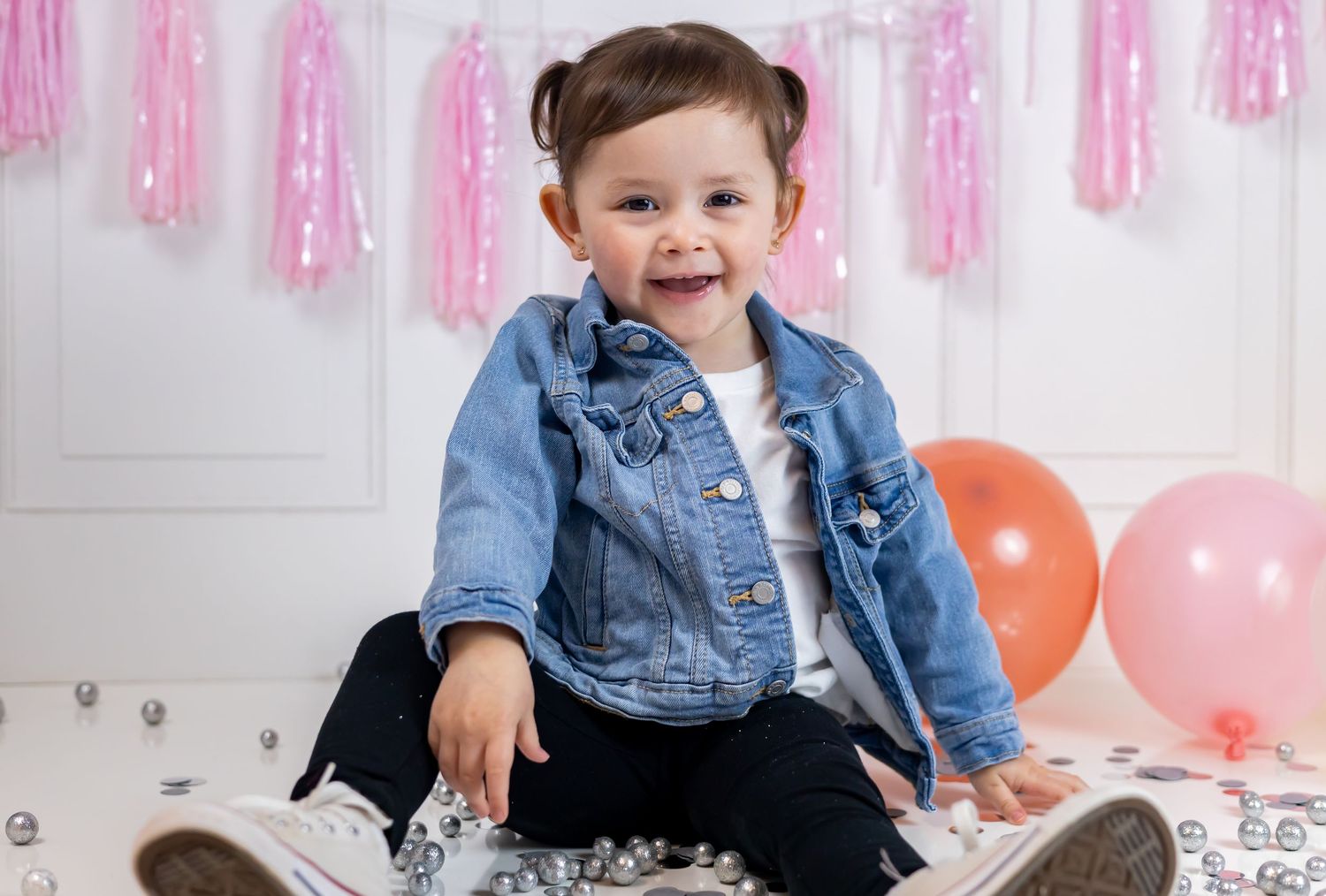 A birthday party scene with pink decorations, colorful balloons, and a cake on a white table against a white backdrop.
