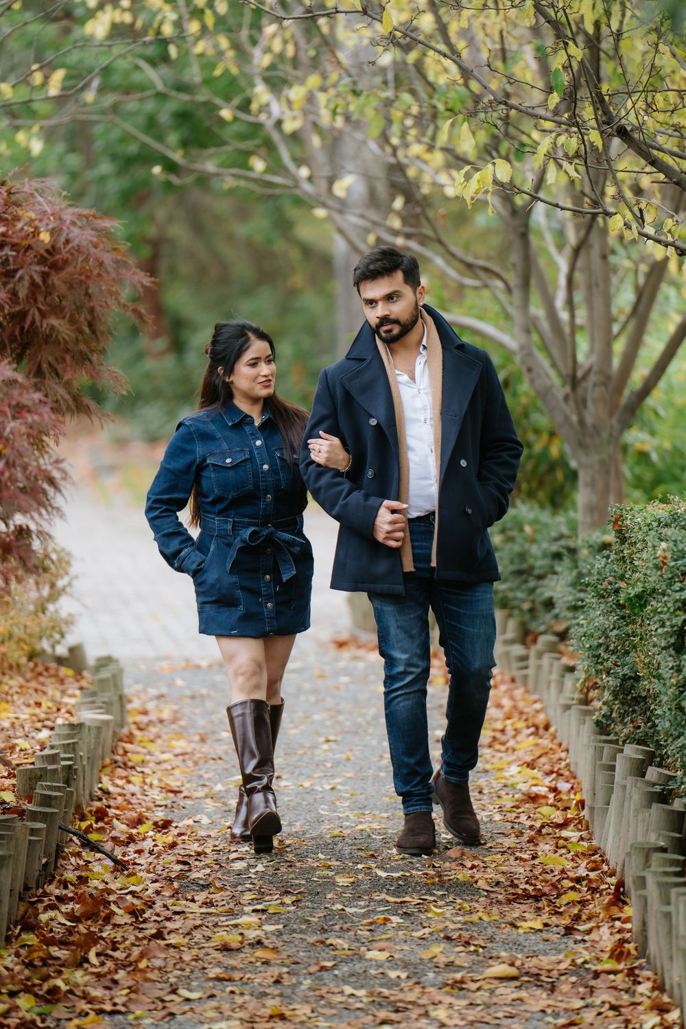 A couple walks together on a leaf-covered path through an autumn park wearing matching denim jackets.