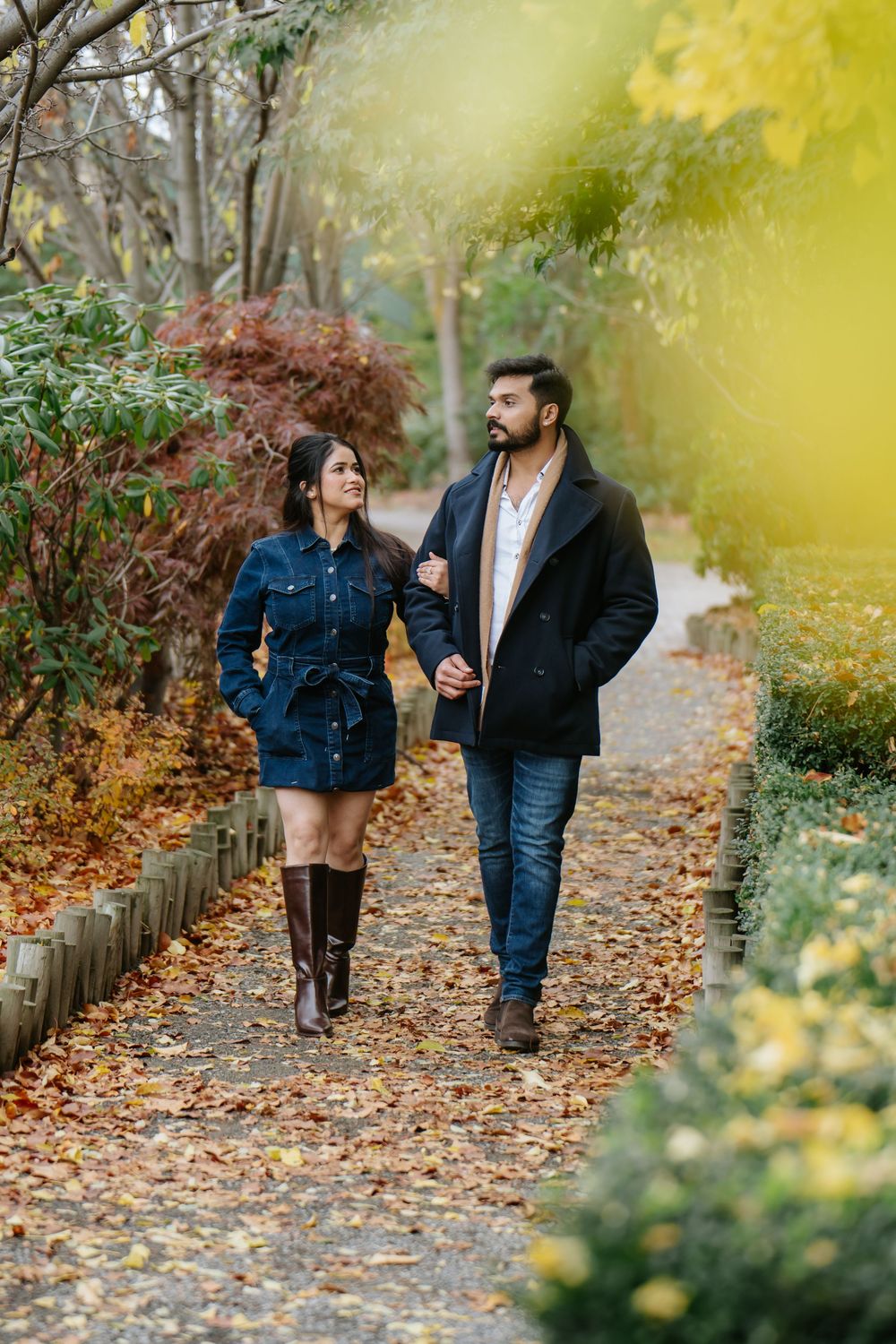 A couple walks together on a leaf-covered autumn path surrounded by yellow foliage and wooden fencing.