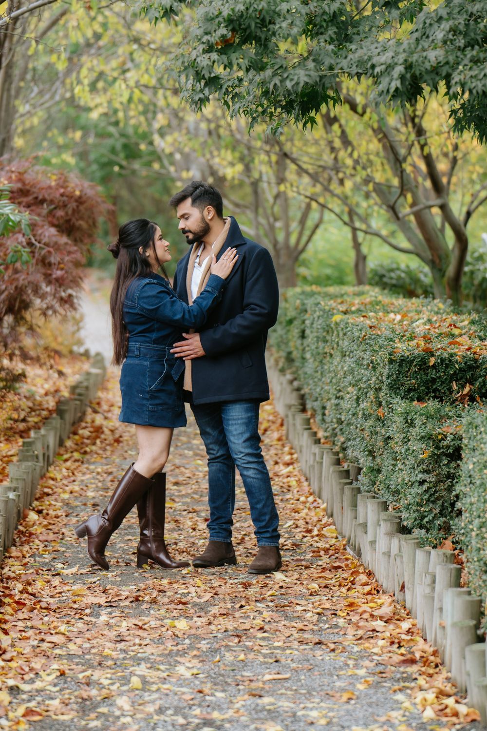 A couple shares romantic moments while walking along a leaf-covered path in an autumn garden setting.