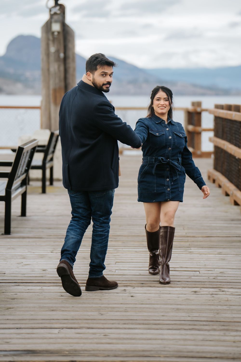 A couple shares a playful moment while walking on a wooden pier with mountains and water in the background.