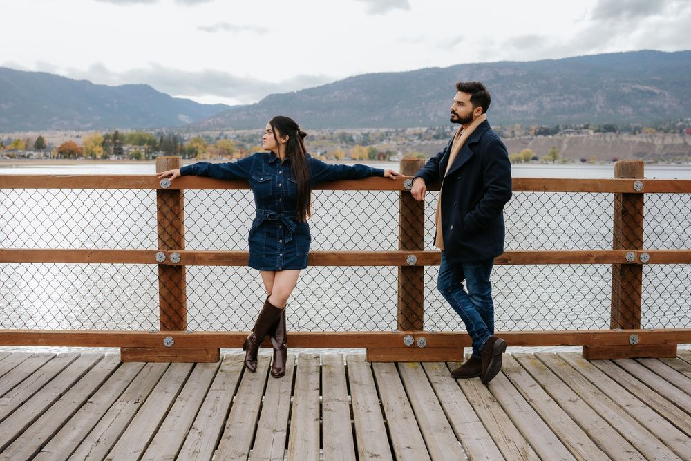 A couple stands together on a wooden dock overlooking a scenic mountain lake on a cloudy day.