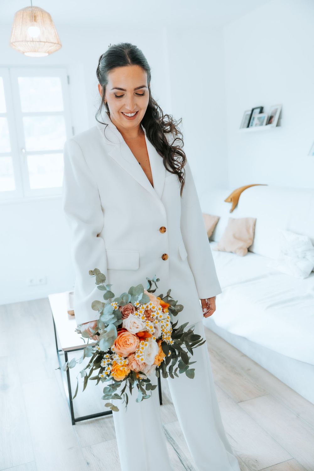 A figure in a white dress holds a colorful bouquet of flowers in a bright room.