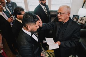 Priest placing hand on groom's head in a pre-wedding blessing ceremony at home.