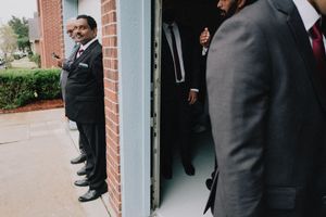 Formally dressed men in suits standing near brick building doorway before wedding ceremony.