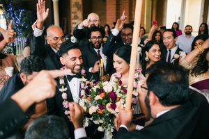 Joyful multicultural wedding couple surrounded by celebrating guests holding colorful floral bouquet.