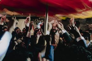 Energetic wedding reception crowd dancing under vibrant red fabric with hands raised in celebration.