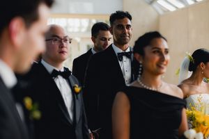 Groom in black tuxedo smiling during wedding ceremony surrounded by wedding party members.
