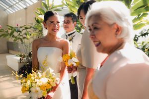 Radiant bride smiling with yellow and white bouquet greeted by elderly woman in greenhouse venue.
